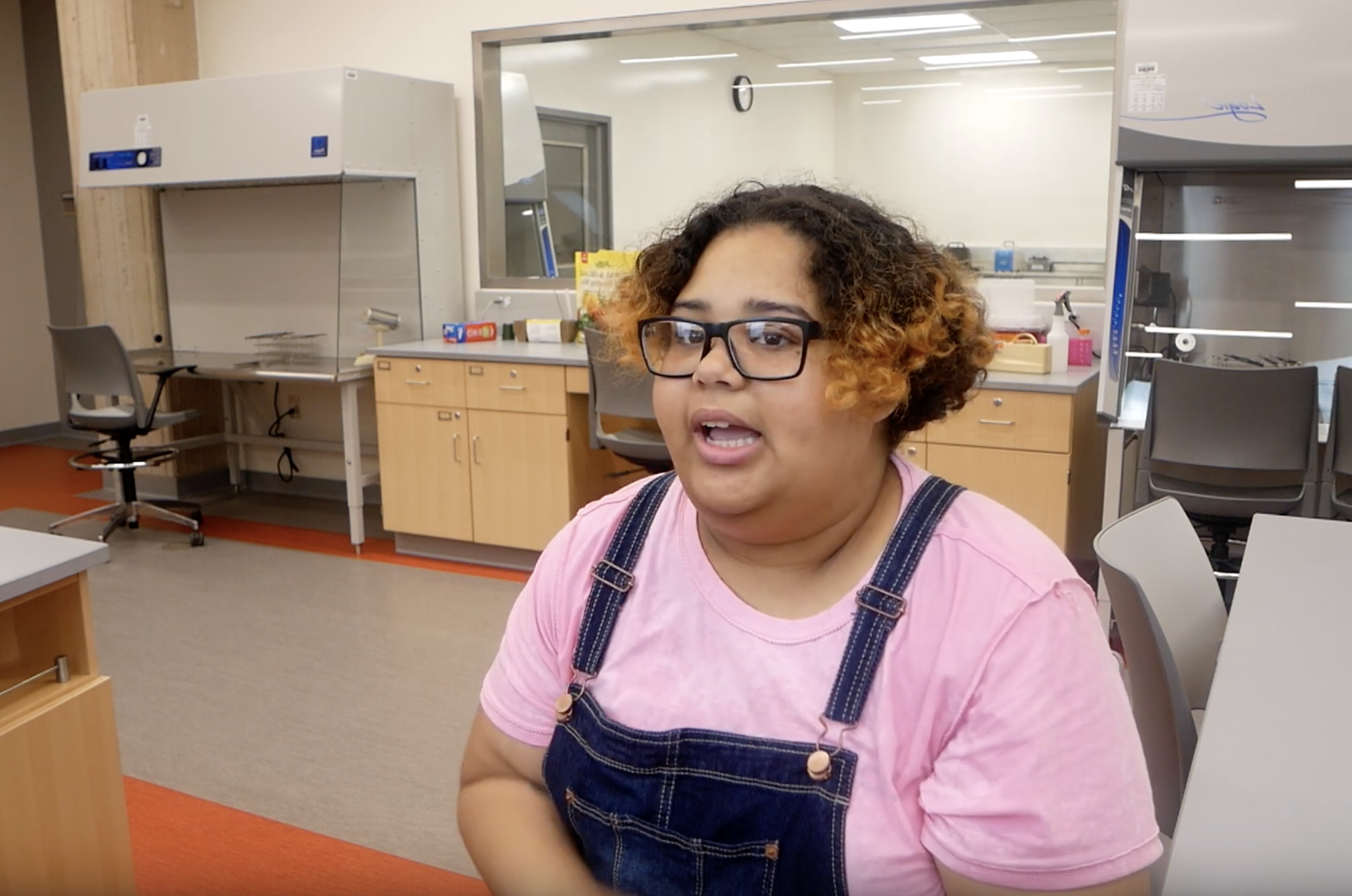 凯什拉·卡布雷拉在实验室讲话的照片. 她穿着粉红色的t恤和蓝色的工作服，戴着眼镜.
